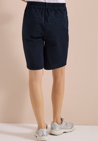 Persona con pantalones cortos negros con cintura elástica y zapatos deportivos grises, de pie sobre un fondo beige.