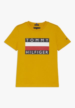 T-shirt in cotone giallo con un logo rettangolare nero, bianco e rosso che presenta la scritta "TOMMY HILFGER". Maniche corte e scollo rotondo.