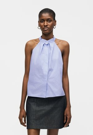 Femeie îmbrăcată într-un top albastru deschis fără mâneci, cu guler cu nasturi, și fustă gri închis, stând în fața unui fundal simplu.