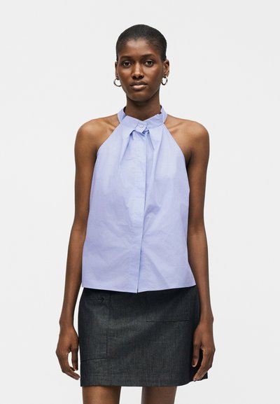 Femme portant un top bleu clair sans manches à col halter avec boutons au niveau du cou et une jupe gris foncé, debout devant un fond uni.