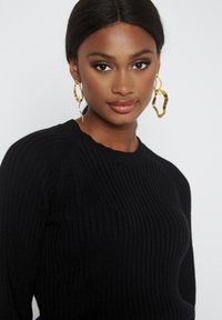 Maglione nero a coste con scollatura arrotondata. Il modello indossa grandi orecchini ad anello astratti in oro, con superficie texturizzata e design moderno.