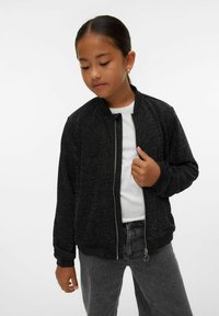 Młoda dziewczyna z ciemnymi włosami związanymi w kucyk, nosząca złote kolczyki, czarną kurtkę z zamkiem, białą koszulę i szare dżinsy na jednolitym białym tle.