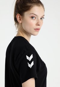 Young woman with light skin and hair tied back, wearing a black t-shirt with white chevron stripes on the sleeve, looking over her shoulder.