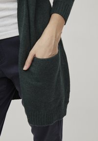 Cárdigan verde oscuro hecho de punto texturizado con puños acanalados y un gran bolsillo lateral, llevado sobre una camisa clara y pantalones oscuros.