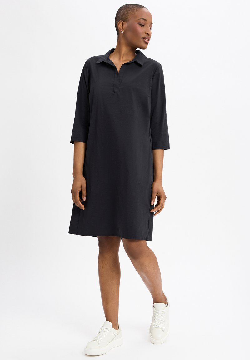 Zwart gekraagde jurk met driekwartmouwen, knielange snit, gemaakt van gladde stof, gecombineerd met witte sneakers.