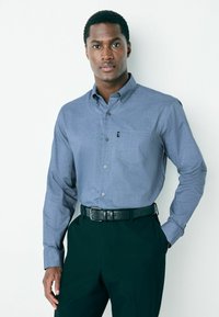 Világoskék anyagból készült, enyhén texturált, gombos ing egy mellzsebbel, sötét nadrággal és fekete övvel párosítva.