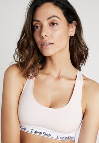Šviesiai rožinė Calvin Klein braletė su apvaliu iškirpimu, plačiomis petnešėlėmis ir kontrastinga elastinga juosta, ant kurios yra prekės ženklo logotipas.