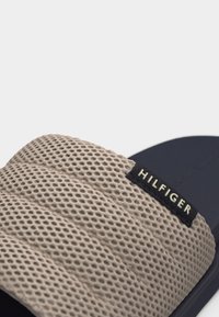 Κοντινό πλάνο ενός σαγιοναριού σε σκούρο μπλε ναυτικό χρώμα και μπεζ με ραβδωτό λουρί από δίχτυ, με την ένδειξη "Hilfiger".