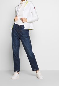 Vitt vadderat zip-up-jacka med huva, parat med mörkblå vida jeans. Vita sneakers kompletterar outfiten.