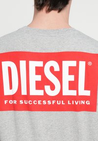 Sivé tričko s červeným obdĺžnikovým grafickým motívom. Motív obsahuje slovo "DIESEL" veľkými bielymi písmenami a pod ním frázu "PRE ÚSPEŠNÝ ŽIVOT".