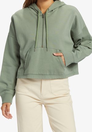 Personne portant un sweat à capuche court vert sauge avec fermeture éclair et poche à l'avant, associé à un pantalon beige clair taille haute, main dans la poche.
