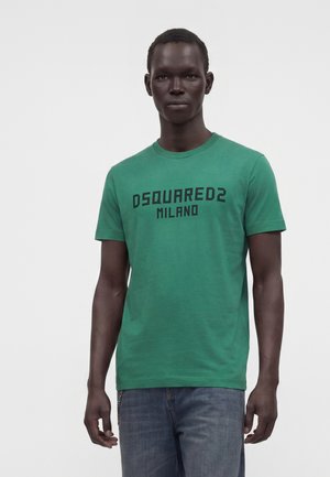 Tânăr purtând un tricou verde cu textul "DSQUARED2 MILANO" și blugi albaștri, stând pe un fundal simplu și luminos.
