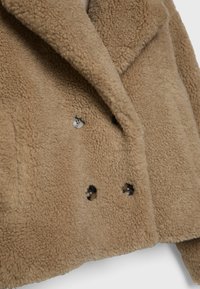 Manteau teddy brun à double boutonnage avec grands revers et quatre gros boutons foncés, mettant en valeur un tissu doux et texturé en fausse toison.