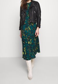 Zwarte leren bikerjack met geribbelde details over een teal bloemenjurk met gele en groene bladeren, gecombineerd met witte cowboylaarzen.