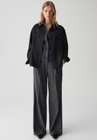 Chemise-jacket noire à boutons avec deux poches poitrine, superposée sur une chemise en denim foncé et un pantalon large gris à rayures, assortie à des talons noirs.