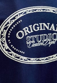 Navyblauw stoffen t-shirt met witte bedrukte tekst en een decoratieve rand. De tekst bevat "ORIGINAL STUDIO" en "Creative Dept."