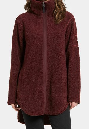 Frau trägt eine bordeauxrote Fleecejacke mit durchgehendem Reißverschluss, hohem Kragen, Textur, langen Ärmeln und leicht geschwungenem Saum über schwarzen Hosen.