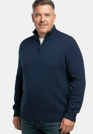 Mężczyzna stoi, ubrany w granatowy sweter na zamek błyskawiczny na białej koszuli i niebieskich dżinsach, neutralne tło.