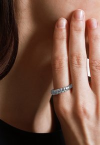 Anello in argento intrecciato su un dito, caratterizzato da un design texturizzato e a spirale. Lo sfondo è sfocato, mettendo a fuoco l'anello e la mano. Pelle liscia visibile.