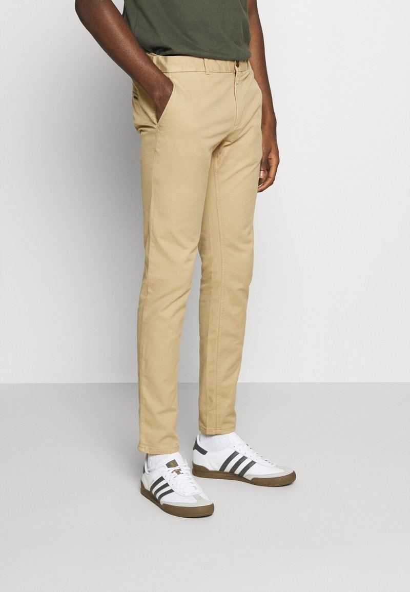 Beige chinos with a slim fit, featuring side pockets and a smooth texture. Paired with white sneakers and a dark green t-shirt.