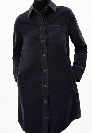 Personne portant une robe-chemise longue en jean noir avec boutons-pression, poche poitrine, manches longues et les mains dans les poches latérales.