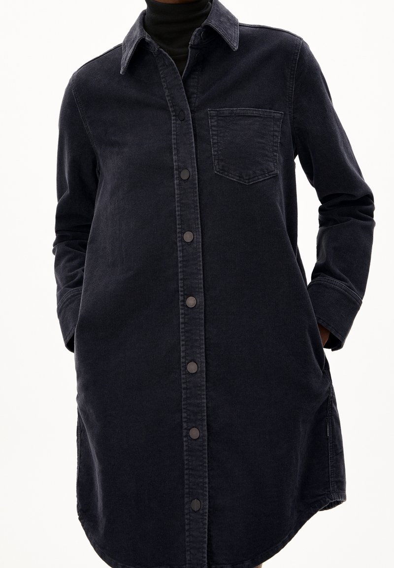 Personne portant une robe-chemise longue en jean noir avec boutons-pression, poche poitrine, manches longues et les mains dans les poches latérales.