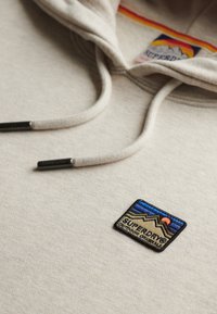 Felpa con cappuccio color beige chiaro realizzata in tessuto morbido, caratterizzata da un cordino con punte metalliche e una toppa ricamata che raffigura montagne e un tramonto.