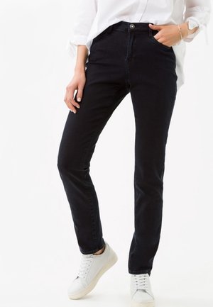 Kvinde iført sorte slim-fit jeans, hvide sneakers og en hvid langærmet skjorte med den ene hånd i lommen og den anden hvilede ved siden af sig.