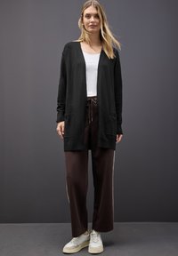 Svart kofta med långa ärmar och fickor fram, matchad med bruna vida byxor och vita sneakers, visad mot en grå bakgrund.