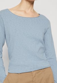 Světle modrý dlouhý rukáv s širokým, zaobleným výstřihem a uvolněným střihem, vyrobený z měkké texturované tkaniny. Kombinovaný s béžovými kalhotami.