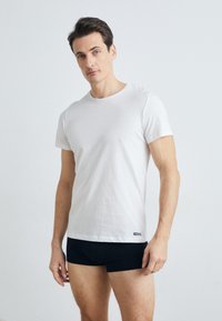 T-shirt blanche à manches courtes en tissu de coton doux, avec un col rond et une petite étiquette logo noire en bas, associée à un short noir.