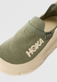 Green slip-on shoe with textured knit upper, cushioned cream sole, and HOKA logo. Soft material and elasticated detail for easy wear.