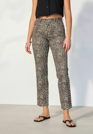 Nainen, jolla on päällään leopardikuosiset suorat housut, musta napitettu lyhyt yläosa, mustat varvastossut ja kultainen rannekoru, seisoo harmaalla lattialla.