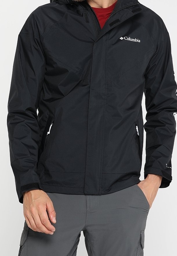 Personne portant une veste noire Columbia avec des poches zippées, un col, et un pantalon cargo gris avec les mains dans les poches.