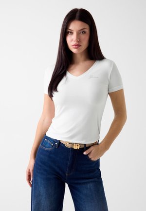 Femme aux longs cheveux foncés portant un t-shirt blanc à col en V et un jean bleu avec une ceinture beige, posant devant un fond clair uni.