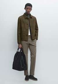 Olijfgroene suède jas, beige broek, donkere suède schoenen en een zwarte stoffen draagtas met schouderbanden, staande tegen een lichte achtergrond.