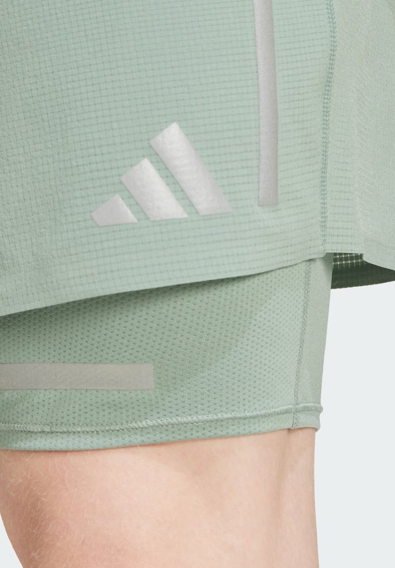 Nahaufnahme von hellgrünen, strukturierten Sportshorts mit Adidas-Logo und reflektierenden Streifen auf einem nackten Bein.