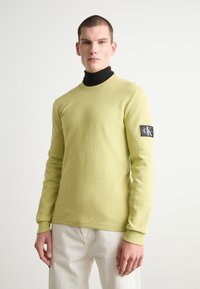 Langermet, teksturert limegrønn skjorte med rund hals og en svart genser med høy hals under. Har et logo-patch på venstre erme.