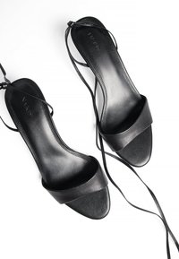 Sandales à talons noirs avec une tige en cuir lisse, bout ouvert et fines sangles autour de la cheville. Le design comprend une large sangle avant et un matériel minimal.