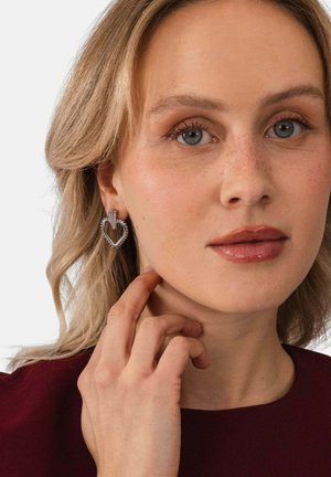 Frau mit blondem Haar, die silberne herzförmige Ohrringe und ein dunkelrotes Oberteil trägt, berührt sanft ihren Hals mit der Hand.