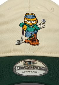 Gorra ajustable con una corona beige y visera verde. Presenta un personaje de dibujos animados bordado jugando al golf, con detalles coloridos en la ropa y accesorios.