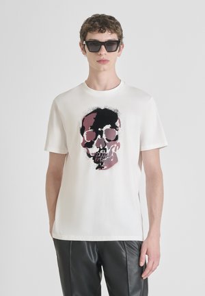 Camiseta blanca de algodón con un gran gráfico de calavera en negro y rosa, cuello redondo y mangas cortas. Llevada con pantalones de cuero negros.