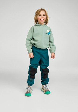 Lächendes Kind mit mintgrünem Hoodie, petrolfarbenen Hosen mit schwarzen Knieaufnähern und beigen Turnschuhen mit grünen Sohlen, stehend vor einem einfarbigen Hintergrund.