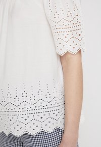 Blusa blanca bordada con patrones recortados en el dobladillo y las mangas, con un borde ondulado. Llevada sobre pantalones de cuadros vichy azules.