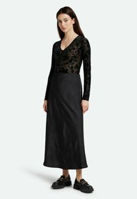 Schwarzes Langarmoberteil mit floraler Spitzenüberlage und V-Ausschnittdesign. Fließender schwarzer Rock mit strukturiertem Muster. Klobige schwarze Schuhe mit Schnallen.