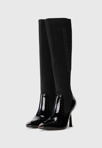 Black high-heeled boots featuring a glossy patent leather toe and studded accents, with a fitted black knit shaft and embossed text detail.