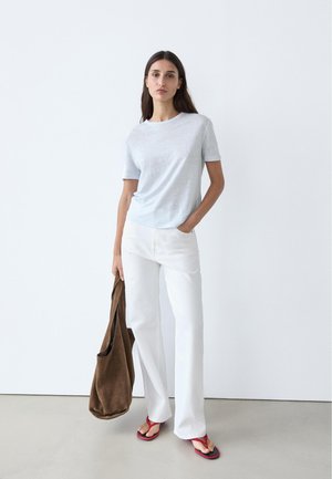 Woman wearing light grey T-shirt, white pants, red flip-flops, holding a large brown tote bag, standing against plain white wall.