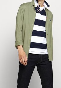 Muž v olivové košili na knoflíky přes námořnickou a bílou pruhovanou ragby polo s tmavými džíny na světlém jednobarevném pozadí.