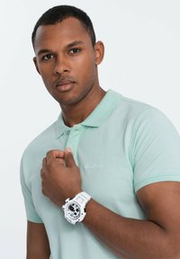 Minttivihreä polo-paita, jossa on pieni brodeerattu logo, yhdistetty valkoiseen kelloon, jonka kellotaulussa on mustaa ja punaista. Sujuva kangas, lyhyet hihat.