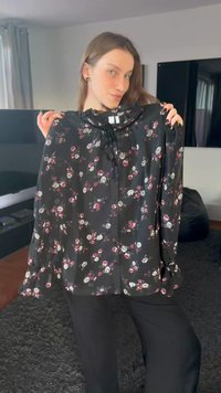 Svart blus med blommönster och långa ärmar, med volangprydd halsringning och små vita och rosa blommor. Materialet verkar vara lätt och flödande.